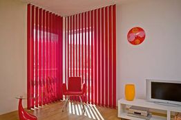 Wohnzimmer mit roten Jalousien, rotem Sessel, Fernseher und dekorativen Elementen in hellen Farben.