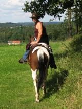 Person auf Pferd, reitet über eine Wiese, trägt Hut, im Hintergrund Hügel und Bäume.