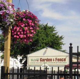 Hanging flower basket near a sign reading "Reliable Garden & Fence" with trees in the background.