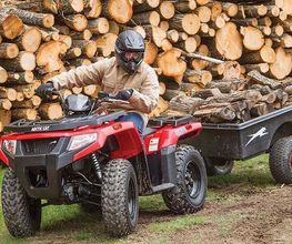 Persona en un ATV rojo traslada leña en un remolque frente a una gran pila de troncos.