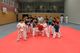 Kinder in Karate-Anzügen machen Kampfposen auf einer roten Matte in einer Turnhalle.