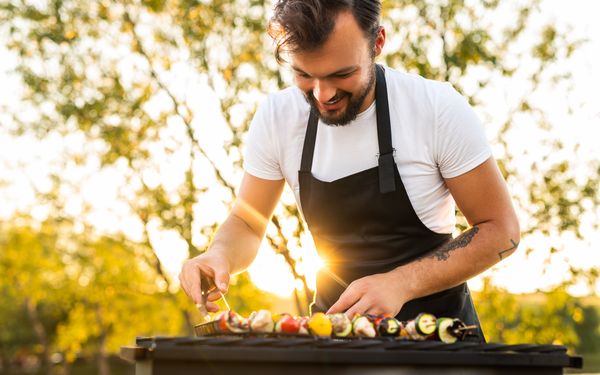 Ein Mann in schwarzer Grillschürze steht vor seinem Grill und grillt Gemüsespiese.