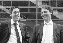 Zwei Männer in Anzügen stehen lächelnd nebeneinander vor moderner Architektur.