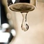 A water droplet hangs from a faucet against a blurred background.