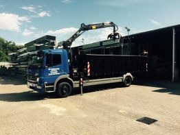 LKW mit Kran lädt Metallteile vor einer Lagerhalle bei sonnigem Wetter ab.