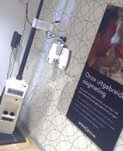 Een oogmeetruimte met een zwarte stoel en een apparaat naast een poster aan de muur.