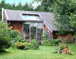 Kleines Holzhaus mit Glasfront, umgeben von Garten und Bäumen.