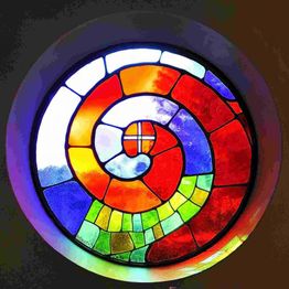 Kreisförmiges Buntglasfenster mit farbigen Mosaiken in Spiralmuster, vorwiegend rot, blau und grün.