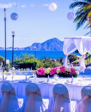 Mesa preparada para evento al aire libre junto al mar, con flores y una pérgola blanca.