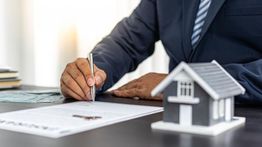 Person signing a document at a desk with a miniature house model beside them.