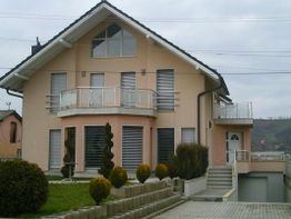 Modernes zweistöckiges Haus mit Balkon und großem Fenster, umgeben von gepflegtem Garten.