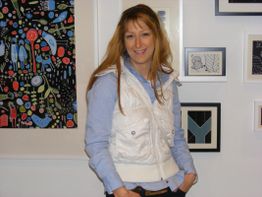 Frau mit weißer Weste steht vor einer Wand mit bunten, gerahmten Bildern.