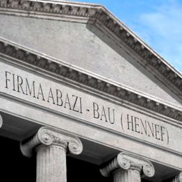 Gebäude mit Säulen und der Inschrift "FIRMA ABAZI-BAU (HENNEF)" oben an der Fassade.