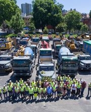 Gruppenfoto von Arbeitern in Warnwesten vor abgestellten Baufahrzeugen auf einem Parkplatz.