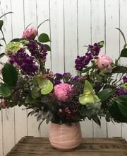 Ramo de flores variadas en jarrón rosado sobre mesa de madera, fondo de pared blanca de madera.