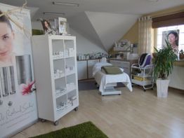 Kosmetikstudio mit Bett, Regal voller Produkte, Pflanzen und großen Werbepostern an den Wänden.