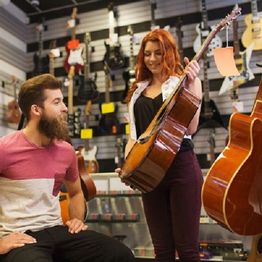 Two people in a music store, looking at guitars with a display of instruments in the background.