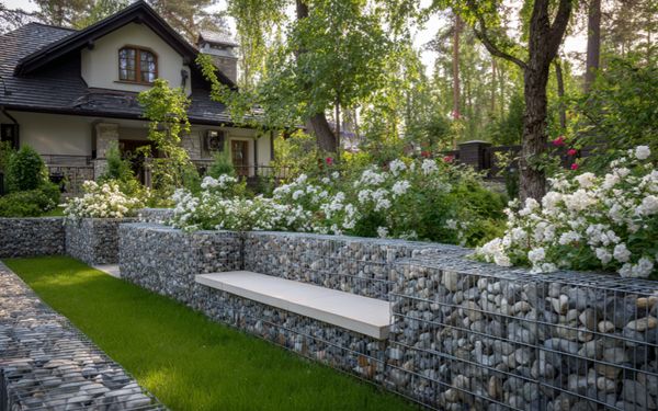 Gabionenzaun mit eingebauter Bank in gepflegtem Garten vor einem Haus.