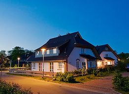 Zwei beleuchtete Häuser am Abend, umgeben von einer Straße und Pflanzen, unter wolkenlosem Himmel.