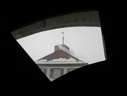 Dach mit Schneedecke und Wetterfahne durch dreieckiges Fenster gesehen.