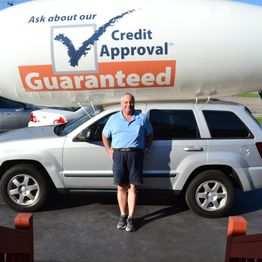 Man in blue shirt stands by an SUV under a "Credit Approval Guaranteed" sign balloon.