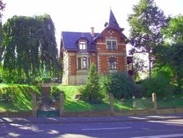 Historisches Backsteinhaus mit Spitzdach, umgeben von Bäumen und grünem Garten, bei sonnigem Wetter.