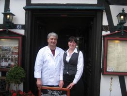 Zwei Personen stehen lächelnd vor einem Fachwerkrestaurant neben einer Tafel.