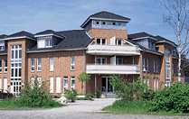 Zweistöckiges rotes Backsteingebäude mit weißen Balkonen und grüner Umgebung.