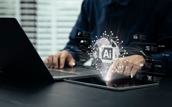 Person arbeitet an Laptop und Tablet mit eingeblendeter KI-Grafik und digitalen Anwendungen.