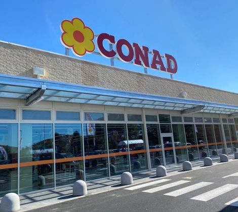 Esterno di un supermercato Conad con insegna rossa e fiore giallo sul tetto.