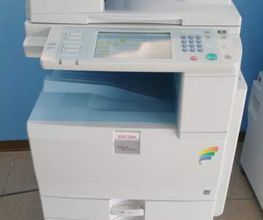 Fotocopiatrice bianca multifunzione su pavimento beige, sfondo azzurro.