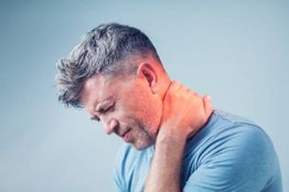 Man with grey hair holds neck in pain, wearing a blue shirt against a plain background.