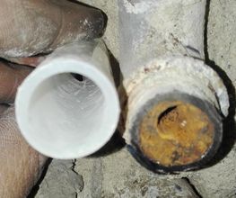 Mano sosteniendo una tubería plástica junto a una tubería de metal oxidada en una pared.
