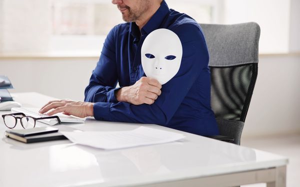 Ein Mann sitzt an einem Schreibtisch und hält sich eine weiße Maske vor seinen Oberkörper, um durch verschleierte Tatsachen einen Versicherungsbetrug zu symbolisieren.