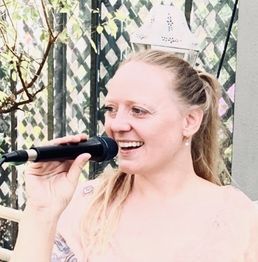 Frau mit Mikrofon lächelt, während sie singt, vor einem geflochtenen Hintergrund im Freien.