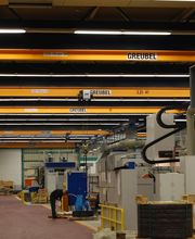 Fabrikhalle mit gelben Brückenkranen, Maschinen und gestapelten Materialien auf Paletten.