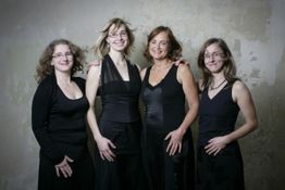 Vier lächelnde Frauen in schwarzen Outfits stehen vor einer beigen Wand.