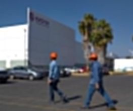 Dos personas con cascos naranjas caminan frente a un edificio blanco y palmeras.