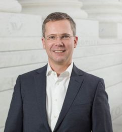 Ein lächelnder Mann in Anzug und Brille steht vor einer weißen Wand mit Schriftzügen.