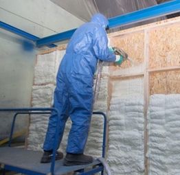 Worker in blue suit spraying foam insulation on a wooden wall.