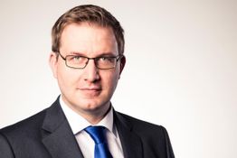 Ein Mann mit Brille im Anzug und blauer Krawatte vor hellem Hintergrund.