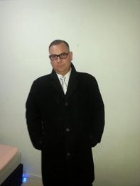 Person im schwarzen Mantel und Krawatte, vor hellgrüner Wand stehend, Hände in den Taschen.