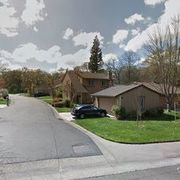 Suburban street with houses, green lawns, trees, and a parked car under a partly cloudy sky.
