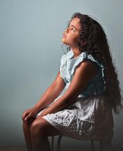 Niña sentada con vestido azul claro, mirando hacia arriba, fondo gris.