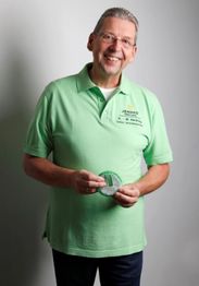 Ein Mann im grünen Hemd lächelt und hält einen runden Gegenstand vor grauem Hintergrund.