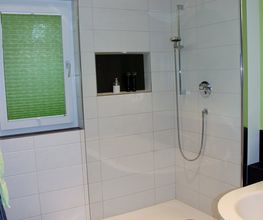 Moderne Dusche mit Glaswänden, weiß gefliest, neben einem Fenster mit grünem Sichtschutz.