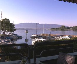 Blick auf einen ruhigen Hafen mit Booten und Bergen im Hintergrund, aufgenommen von einer Terrasse.