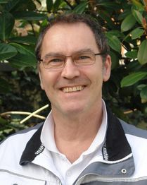 Ein lächelnder Mann mit Brille steht vor grünen Blättern.