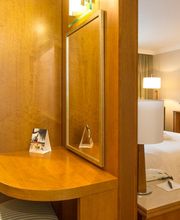 Quarto de hotel com cama, TV, mesa com espelho e cadeiras. Cortinas compridas ao fundo.