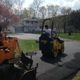 A person operates a roller to pave a driveway near a house surrounded by trees.